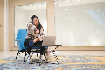 Young Asian girl making a video call on a laptop with her thumb down. The young woman is sitting in a shopping mall.