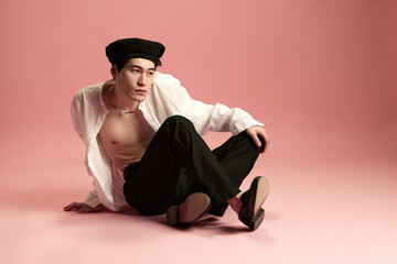 Portrait of stylish, young, korean guy, posing on floor in classical white blouse, pants and black beret against pink studio background. Concept of men's fashion, style, art, beauty, trends