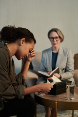 Young depressed female patient touching head during psychological session while sitting against professional counselor listening to her