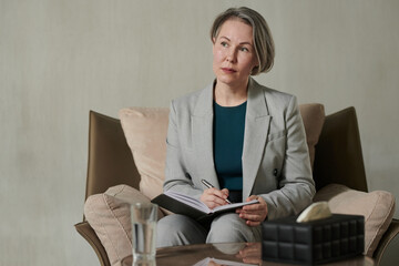Pensive middle aged psychologist with pen over page of open notebook sitting in armchair and...