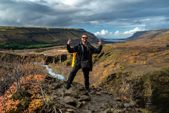 hiker with backpack with victory sign on cliff at the glymur canyon edge in iceland with river and low clouds on background