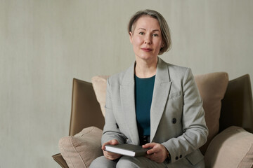 Successful mature psychologist wearing grey elegant suit sitting in armchair against grey wall in office and looking at camera - obrazy, fototapety, plakaty