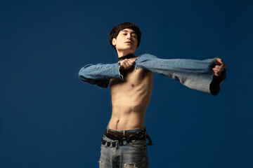 Portrait of young man posing shirtless with jeans sleeve elements against blue studio background....