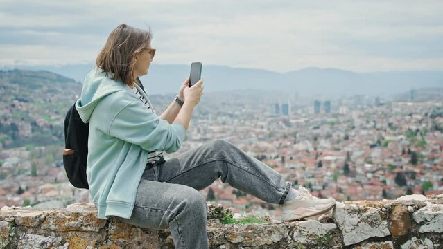 A young adult Caucasian woman taking pictures of the city of Sarajevo from a viewpoint with a smartphone while traveling to the Balkans.