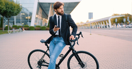 Handsome confident positive young bearded man standing near his bike using phone on the city...