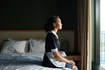Young tired chambermaid in uniform sitting on double bed with two pillows in hotel room and looking...