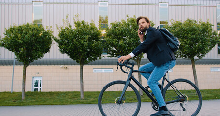 Side view of handsome smiling young bearded man in stylish clothes which riding a bike and has positive phone conversation