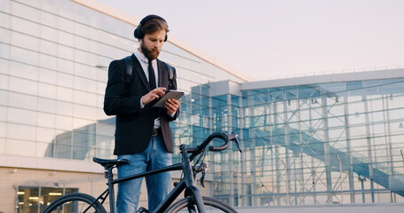 Portrait of handsome concentrated young bearded businessman in earphones which standing near his...