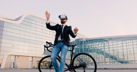 Attractive confident purposeful 30-aged bearded business man in virtual reality headset standing near his bicycle and working on imaginary screen on the background of modern city building