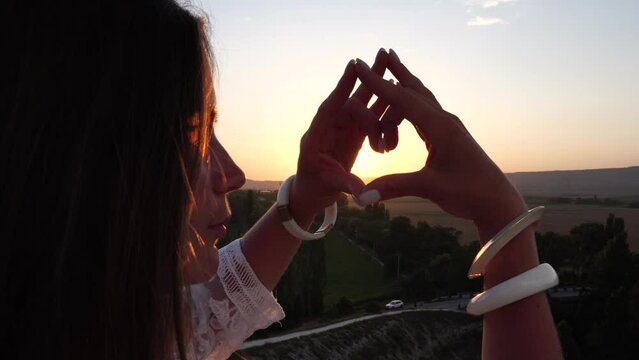 Happy woman in white boho dress making heart sign with hands on sunset in mountains. Romantic woman with long hair standing with her back on the sunset in nature in summer with open hands.
