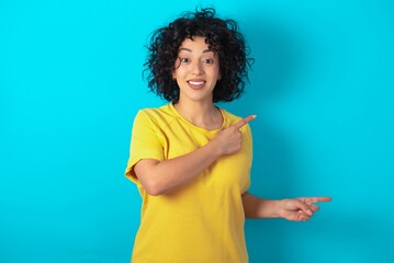 Optimistic Young arab woman wearing yello T-shirt over blue background points with both hands and  looking at empty space.