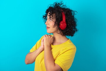 Young arab woman wearing yello T-shirt over blue background wears stereo headphones listening to...