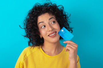Lovely Young arab woman wearing yello T-shirt over blue background showing credit card and looking...
