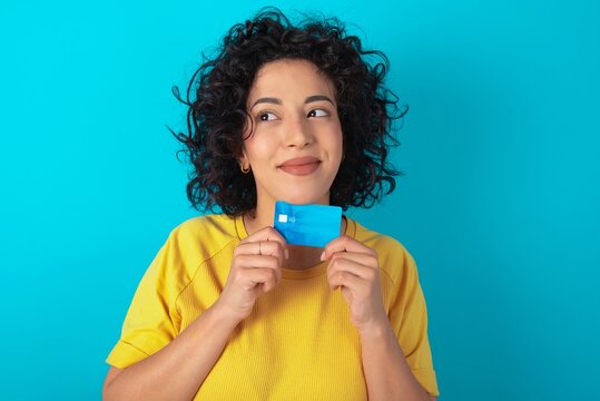 Photo of cheerful Young arab woman wearing yello T-shirt over blue background hold debit card look empty space