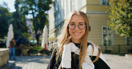 Fototapeta premium Good-looking happy joyful modern young blondie in glasses looking at camera with sincerely smile on the background of ancient city building on sunny day