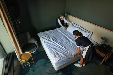 Above angle of young chambermaid and manager preparing double bed for guests while changing...