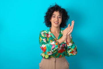 young arab woman wearing colorful shirt over blue background clapping and applauding happy and...