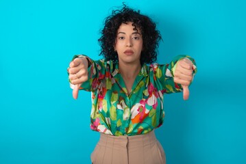 young arab woman wearing colorful shirt over blue background being upset showing thumb down with...