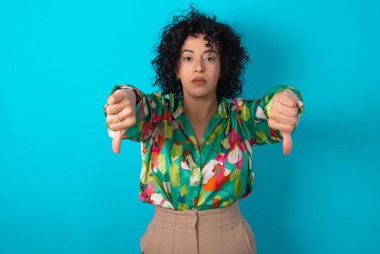 young arab woman wearing colorful shirt over blue background being upset showing thumb down with two hands. Dislike concept.