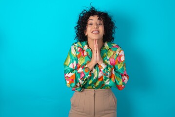 young arab woman wearing colorful shirt over blue background praying with hands together asking for forgiveness smiling confident.