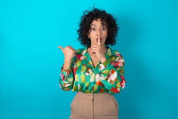 young arab woman wearing colorful shirt over blue background asking to be quiet with finger on lips pointing with hand to the side. Silence and secret concept.