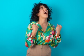 young arab woman wearing colorful shirt over blue background celebrating surprised and amazed for success with arms raised and eyes closed. Winner concept.