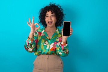 young arab woman wearing colorful shirt over blue background holding in hands cell showing ok-sign