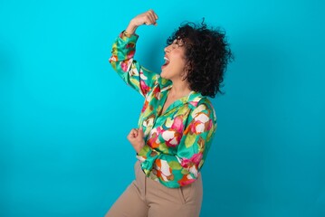 Profile photo of excited young arab woman wearing colorful shirt over blue background good mood raise fists screaming rejoicing overjoyed basketball sports fan supporter
