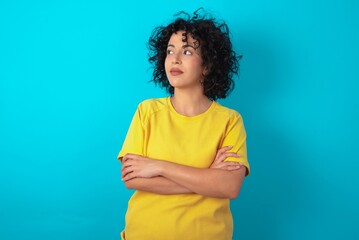 Pleased young arab woman wearing yellow T-shirt over blue background keeps hands crossed over chest...