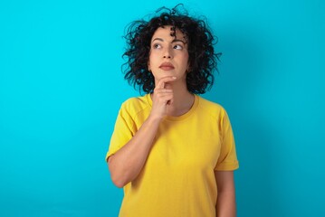 Thoughtful young arab woman wearing yellow T-shirt over blue background holds chin and looks away pensively makes up great plan