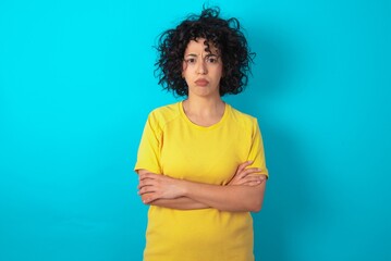 Gloomy dissatisfied young arab woman wearing yellow T-shirt over blue background looks with...