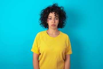 Fototapeta na wymiar Offended dissatisfied young arab woman wearing yellow T-shirt over blue background with moody displeased expression at camera being disappointed by something