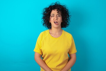 Portrait of dissatisfied young arab woman wearing yellow T-shirt over blue background smirks face,...