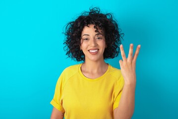 young arab woman wearing yellow T-shirt over blue background smiling and looking friendly, showing...