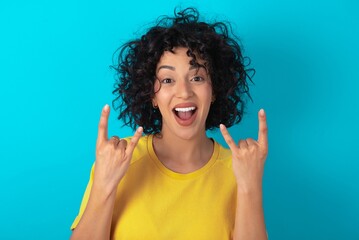 young arab woman wearing yellow T-shirt over blue background makes rock n roll sign looks self confident and cheerful enjoys cool music at party. Body language concept.