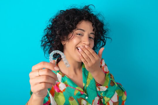 Happy young arab woman wearing colorful shirt over blue background holding and showing at camera an invisible aligner while laughing. Dental healthcare and confidence concept.