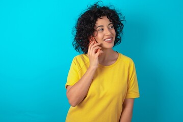 Fototapeta na wymiar Pleasant looking happy young arab woman wearing yellow T-shirt over blue background has nice telephone conversation and looks aside, has nice mood and smiles positively while talks via cell phone