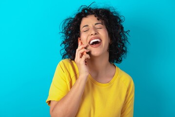 Funny young arab woman wearing yellow T-shirt over blue background laughs happily, has phone conversation, being amused by friend, closes eyes.
