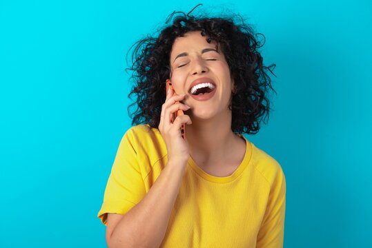 Funny young arab woman wearing yellow T-shirt over blue background laughs happily, has phone conversation, being amused by friend, closes eyes.