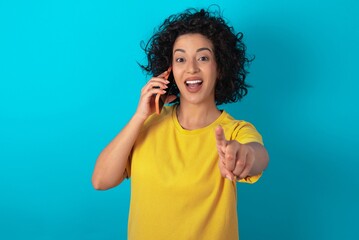Positive young arab woman wearing yellow T-shirt over blue background indicates directly at camera...