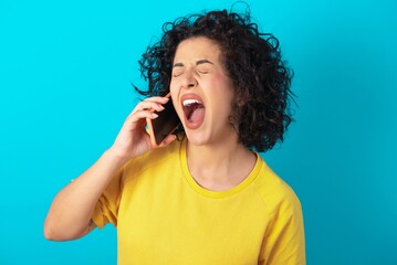 Overemotive happy young arab woman wearing yellow T-shirt over blue background laughs out positively hears funny story from friend during telephone conversation