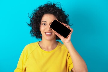 young arab woman wearing yellow T-shirt over blue background holding modern smartphone covering one eye while smiling