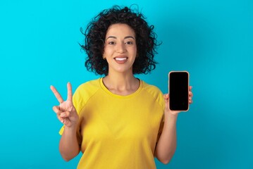 young arab woman wearing yellow T-shirt over blue background holding modern device showing v-sign