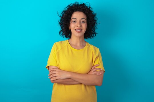 Portrait of charming young arab woman wearing yellow T-shirt over blue background standing confidently smiling toothily with hands folded