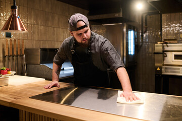 Young male chef in uniform wiping stove surface with clean duster while bending over large wooden...