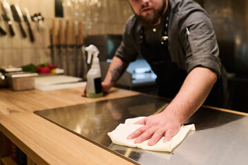 Close-up of young professional chef sanitizing and cleaning surface of electric stove before...