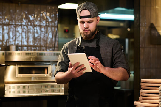 Male cook looking through online recipes on tablet screen while standing by workplace in the kitchen before preparing pizza for guests