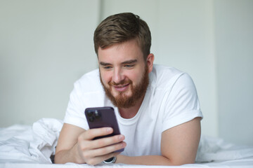 Young bearded happy man is using cell mobile phone at home in bed in bedroom, looking at screen of smartphone 