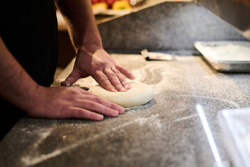 Close-up of hands of young chef kneading in flour fresh dough for pizza on table while preparing traditional italian food in restaurant