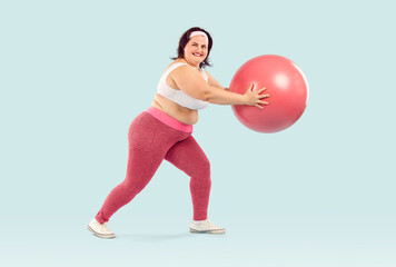 Full length photo of a happy young smiling fat overweight woman wearing sportswear doing exercise with fit ball isolated on studio blue background. Workout sport, fitness and body positive concept.